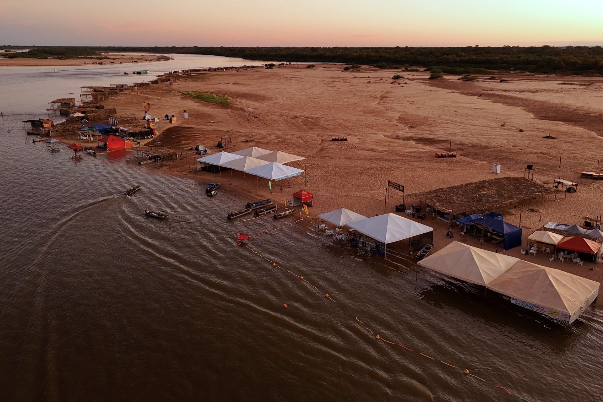 Vista aérea da praia do Rio Araguaia ao pôr do sol, com tendas, barcos e banco de areia às margens do rio